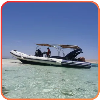 A small inflatable boat with a canopy, moored in shallow, clear turquoise waters, with an operator standing aboard.