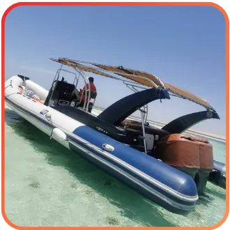 A small inflatable boat with a canopy, moored in shallow, clear turquoise waters, with an operator standing aboard.