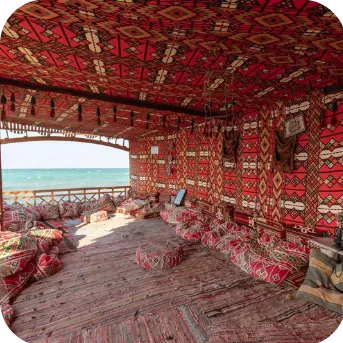 An interior view of a beachside cabana with vibrant red and patterned cushions and rugs, overlooking the sea.