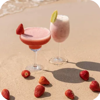 Two refreshing drinks on the sandy beach, one garnished with a strawberry and the other with a lime slice, surrounded by scattered strawberries.