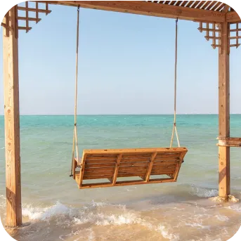 A wooden swing hanging from a pergola over a sandy beach, with waves gently lapping at the swing's base.