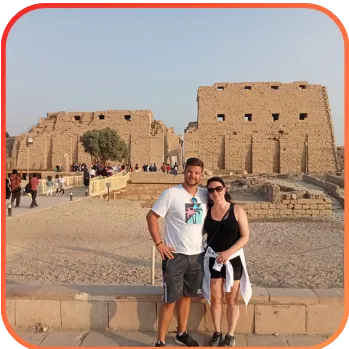 A couple posing in front of a large ancient stone building with rectangular structures, likely tourists at a historic site.