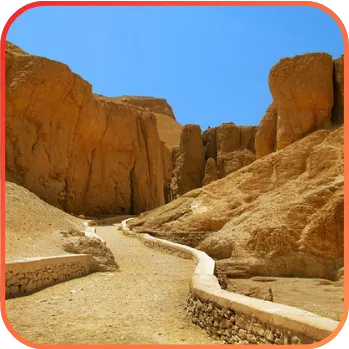 A narrow, winding valley path between tall, sheer rock walls under a bright blue sky, indicative of a desert canyon.