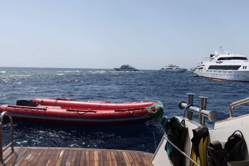 A view from the stern of a boat showing a red inflatable dinghy on the water, with multiple white yachts in the background on a sunny day.